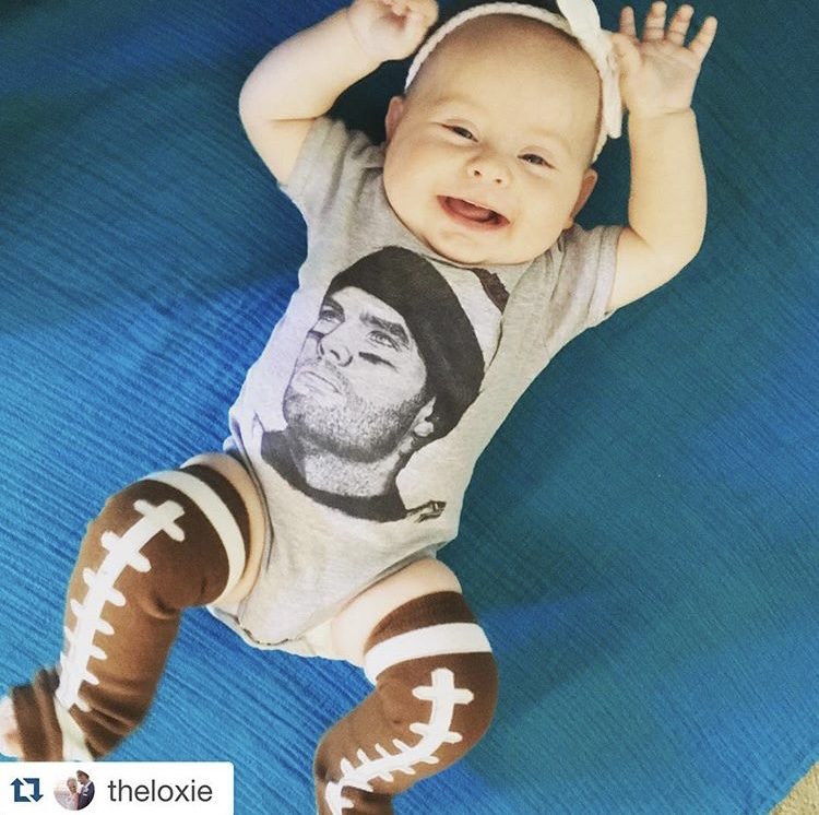A smiling baby in a gray 500 Level Tom Brady onesie and football-striped leg warmers.